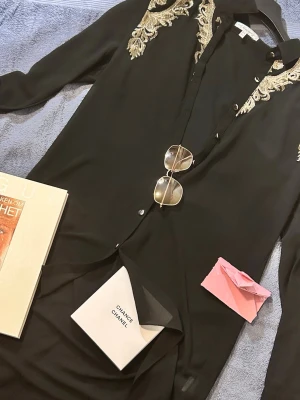 Svart lång skjorta med broderade detaljer - Svart långärmad skjorta från UK med snygga guldiga broderade detaljer vid axlarna. Skjortan har knappar framtill och klassisk krage och är genomskinlig. Perfekt för dig som vill sticka ut med en elegant touch. Vintage 