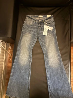 Blå bootcut jeans från Gina Tricot - Ett par blå bootcut jeans från Gina Tricot med klassisk femficksdesign och snygga slitningar framtill. Jeansen är låg midjde och är tillverkade i bomull med en liten mängd elastan för extra komfort. Perfekta för en trendig och avslappnad look.