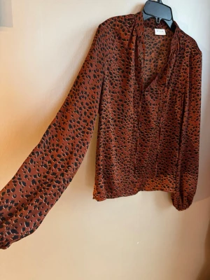 Rostfärgad, leopardmönstrad blus från VILA - Snygg rostfärgad blus från VILA med svart leopardmönster. Blusen har långa ärmar med resår vid ärmslut och en lös passform. Fin knytning i halsringningen. Mycket användbar!