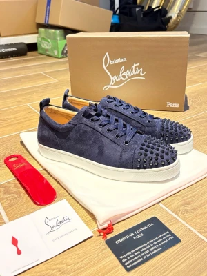 Blå Christian Louboutin sneakers med nitar - Mörkblå sneakers i mocka från Christian Louboutin med svarta nitar på tån och klassisk röd sula. Skorna har lågt skaft, matchande blå skosnören och vita gummisulor. Perfekt för dig som vill sticka ut med exklusiv design och edgy detaljer.