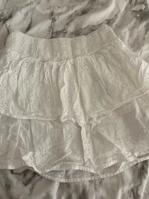 Vit volangkjol från Gina Tricot - Supersöt vit kortkjol från Gina Tricot med broderade hålmönster och volanger. Kjolen har en bred, smockad midja som sitter bekvämt och ger en snygg passform. Perfekt för varma dagar och somriga outfits!