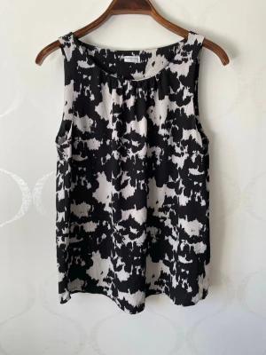 Svartvit ärmlös blus med mönster - Snygg ärmlös blus i svart och vitt med abstrakt mönster. Blusen har rund halsringning och ett avslappnat, rakt fall. Perfekt att matcha med jeans eller kjol för en trendig look.