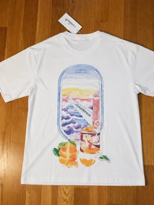  Casablanca t-shirt - Vit t-shirt från Casablanca med färgglatt tryck på ryggen som föreställer ett flygplansfönster, apelsiner och ett glas juice. Mindre tryck på bröstet med texten 'Casa Airlines'. Klassisk passform och rund hals. Perfekt för dig som gillar unika prints.