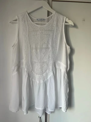 Vit ärmlös blus med spetsdetaljer - Superfin vit blus från Mangomed broderade spetsdetaljer framtill. Modellen är ärmlös och har en lös, flowy passform som ger en bohemisk vibe. Perfekt att styla med jeans eller shorts för en fräsch look.