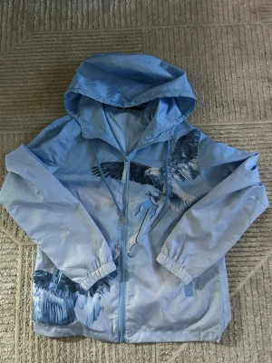 Blå vindjacka med fågeltryck Trapstar - Ljusblå vindjacka från Trapstar med huva och dragkedja. Jackan har ett stort tryck av flygande fåglar både fram och bak samt Trapstar-logga på ryggen. Tillverkad i lätt, vindtätt material och har resår vid ärmsluten. Perfekt för en streetwear-look.