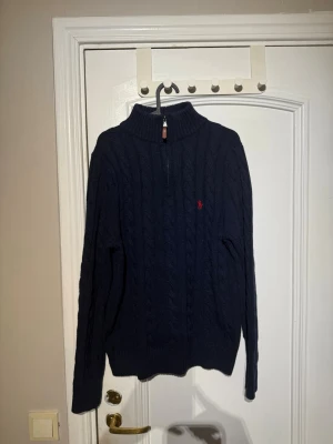 Mörkblå kabelstickad tröja Polo Ralph Lauren - Mörkblå kabelstickad tröja från Polo Ralph Lauren med halv dragkedja och hög krage. Tröjan har det klassiska röda broderade logotypen på bröstet och ribbade muddar vid ärmslut och nederkant. Perfekt för lager-på-lager-stil.