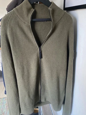 Olivgrön stickad cardigan J.Lindeberg - Säljer en olivgrön stickad cardigan  från J.Lindeberg med dragkedja framtill och hög krage. Tröjan har ribbstickad struktur och långa ärmar, perfekt för lager på lager. Klassisk och stilren design som passar till mycket.