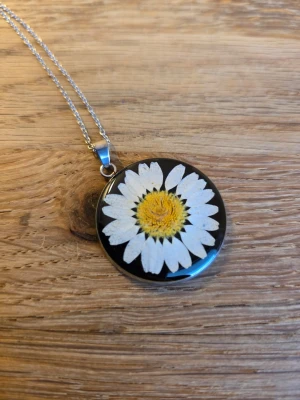 Halsband med prästkrage - Unikt halsband med en riktig prästkrage inbäddad i resin. Hängsmycket är runt med svart bakgrund och har en silverfärgad kedja. Perfekt accessoar för dig som gillar naturinspirerade smycken och vill sticka ut med en blomdetalj.