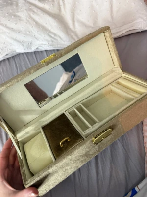 Guldig smyckesbox med spegel - Snygg smyckesbox i guldig sammet med spegel på insidan av locket. Flera fack för ringar, örhängen och armband samt ett litet handtag i sammet. Låset är i guldfärgad metall och boxen har en lyxig känsla. Perfekt för att organisera dina accessoarer.