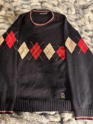 Mörkblå argyletröja från Emilio Cloud - Stickad mörkblå tröja med klassiskt argylemönster i rött, vitt och beige över bröstet. Rund halsringning med randig detalj och ribbstickade muddar. Liten broderad logga nertill. Perfekt för en preppy look.