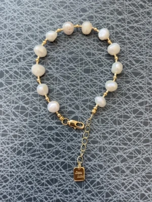 Guldarmband med vita pärlor från Sassy - Stilrent armband med oregelbundna vita pärlor och guldfärgade detaljer. Armbandet har en justerbar kedja och en liten bricka med Sassy-loggan. Perfekt accessoar för att lyfta din outfit med en elegant touch.