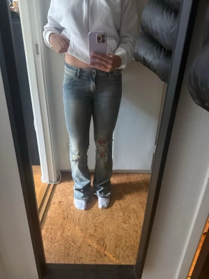 Blå slitna bootcut jeans - Säljer ett par blå bootcut jeans med slitningar och hål på ena knät. Jeansen har klassisk femficksdesign och är i stretchigt denimtyg. Perfekta för en avslappnad och trendig look.