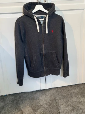 Mörkgrå hoodie från Polo Ralph Lauren - Mörkgrå hoodie med dragkedja från Polo Ralph Lauren. Klassisk design med vit dragsko i huvan, känguruficka och röd broderad logga på bröstet. Perfekt för en avslappnad och stilren look. Sjukt fräsch tröja som knappt är använd. Ställ gärna frågor😃