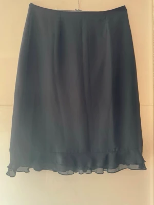 Svart långkjol med volangkant - En stilren svart långkjol med lätt volangkant nertill. Kjolen har en enkel och klassisk design med mjukt fall och är perfekt för dig som gillar en tidlös look. Materialet känns lätt och flowy, vilket ger kjolen en snygg siluett.