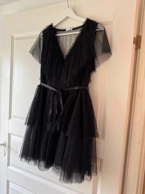 Svart volangklänning med bälte S - Supersnygg svart kortklänning i flera lager tunn tyll med volanger och v-ringning. Klänningen har korta ärmar och ett matchande bälte i midjan som knyts. Perfekt för dig som vill ha en statement piece till garderoben.
