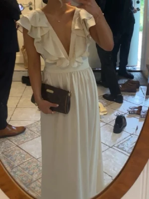 Champagnefärgad långklänning med volang - Elegant champagnefärgad långklänning med djup v-ringning och volangdetaljer. Klänningen har ärmlös design och markerad midja, perfekt för speciella tillfällen som till exempel bal🥰