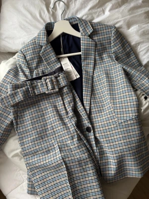 Rutig kostym från Esprit i blått och beige - Snygg tvådelad kostym från Esprit med rutig design i blått, beige och vitt. Kavajen har klassisk krage, enkel knappstängning och två fickor framtill. Byxorna har matchande mönster och bälte i midjan. Perfekt för en stilren look.