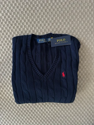 Mörkblå kabelstickad tröja Polo Ralph Lauren - Mörkblå kabelstickad tröja från Polo Ralph Lauren med V-ringning och klassisk röd logga broderad på bröstet. Tröjan har långa ärmar och ribbade muddar. Perfekt för en stilren och tidlös look.