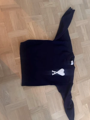 Mörkblå stickad tröja från Ami - Mörkblå stickad tröja från Ami Alexandre Mattiussi med vit hjärtlogga och bokstaven A på bröstet. Tröjan har rund halsringning och ribbstickade muddar vid ärmslut och nederkant. Perfekt för en stilren och trendig look.