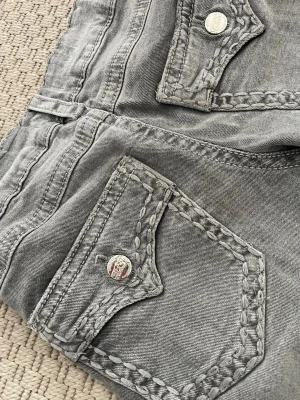 Grå jeans från Gina tricot - Säljer ett par grå jeans med unika, grovt sydda ficklock och dekorativa sömmar. Jeansen har silverfärgade knappar med logga och är tillverkade i klassisk denim. Perfekt för dig som gillar en lite tuffare stil. Har dock en liten fläck (se bild 2)