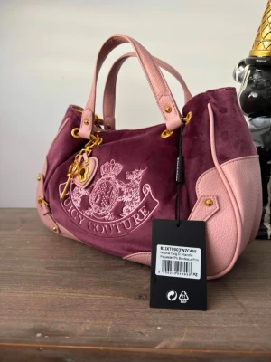 Lila och rosa handväska Juicy Couture - Juicy Couture handväska i lila sammet med ljusrosa detaljer i läder. Väskan har broderad logga framtill, guldfärgade metalldetaljer och både handtag och axelrem. Perfekt för dig som vill sticka ut med en trendig accessoar.