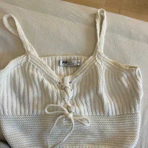 Vit stickad linnetopp - Supersöt vit stickad linnetopp med snörning framtill och smala axelband. Perfekt till sommaren och enkel att styla med jeans eller kjol. Toppen har en ribbad struktur och är croppad i modellen.