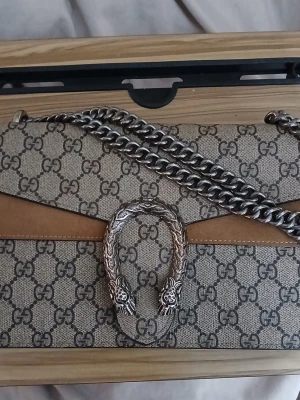 Gucci Dionysus GG Supreme handväska - Snygg handväska från Gucci i beige canvas med klassiskt GG-mönster och bruna mockadetaljer. Väskan har en silverfärgad kedjerem och ikoniskt spänne med två tigrar. Perfekt för dig som vill ha en statementväska med lyxig känsla.