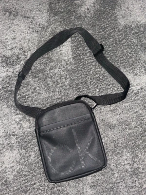 Svart axelväska från Calvin Klein - Snygg svart axelväska från Calvin Klein med justerbar rem och stort CK-logga framtill. Väskan är fyrkantig och tillverkad i slitstarkt syntetmaterial, perfekt för att bära det viktigaste. Enkel och stilren design som passar till många outfits.