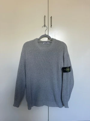 Grå stickad tröja från Stone Island - Snygg grå stickad tröja från Stone Island med klassisk rund halsringning och ribbade muddar. Tröjan har den ikoniska Stone Island-loggan på vänster ärm och är tillverkad i ett grovstickat material som känns både stilrent och bekvämt.