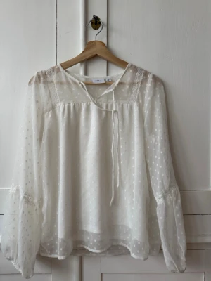 Vit prickig blus från VILA - Söt vit blus från VILA med diskret prickigt mönster och transparenta detaljer. Blusen har långa ärmar med spetsdetaljer och knytband vid halsen. Storlek 34, bra skick ( har en sminkfläck , se bild 3, men går förmodligen bort i tvätt )