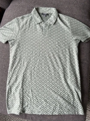 Grönmönstrad pikétröja från SMOG - Säljer en grön pikétröja från SMOG i storlek XS. Tröjan har ett diskret geometriskt mönster över hela plagget, klassisk krage och korta ärmar. Materialet är mjukt och känns som bomull, perfekt för en avslappnad stil.