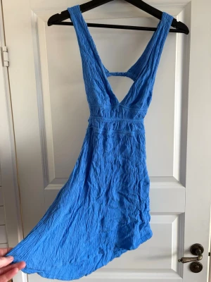 Blå kort klänning med öppen rygg - Snygg blå kortklänning med djup v-ringning och öppen rygg. Klänningen har ett crinklat tyg i lätt material och breda axelband. Perfekt för varma dagar och sommarens äventyr.