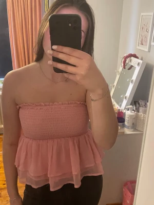 Rosa volangig tubtopp - Söt rosa tubtopp med smockad överdel och dubbla volanger i chiffongliknande material. Toppen är axelbandslös och har en luftig, flowy passform.