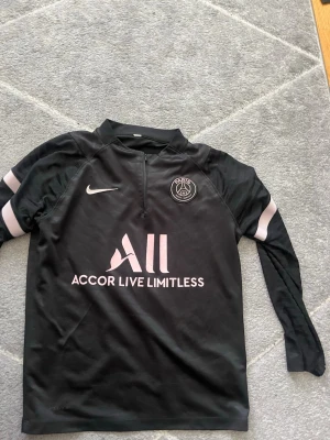 Svart PSG träningströja från Nike - Svart långärmad träningströja från Nike med PSG-logga på bröstet och sponsortryck i vitt och rosa. Tröjan har dragkedja vid halsen och rosa detaljer på ärmarna. Perfekt för fotbollsträning eller sportiga aktiviteter. Det är storlek m men sitter som S