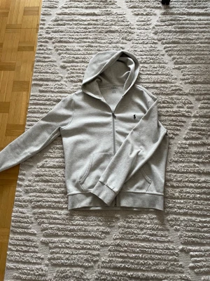Ljusgrå hoodie från Polo Ralph Lauren - Snygg ljusgrå hoodie från Polo Ralph Lauren med dragkedja och klassisk svart logga på bröstet. Hoodien har huva med dragsko och två fickor framtill. Tillverkad i mjuk bomullsblandning som är skön att ha på sig.