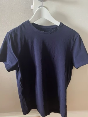 Mörkblå slim fit t-shirt från H&M - En stilren mörkblå t-shirt från H&M i slim fit-modell. T-shirten har rund halsringning och korta ärmar, tillverkad i mjuk bomull som känns skön mot huden. Perfekt för en enkel och clean look.