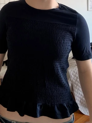 Mörkblå topp med volangkant - Snygg mörkblå topp med korta ärmar och rund halsringning. Toppen har en smockad struktur över hela framsidan och en volangkant längst ner som ger en söt touch. Tillverkad i mjuk bomull, perfekt för en trendig vardagslook. Inga defekter och knappts använts. 