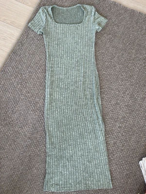Grön ribbad långklänning - En snygg långklänning i ljusgrön nyans med ribbat mönster och korta ärmar. Klänningen har en fyrkantig halsringning och är figurnära. Perfekt för dig som gillar enkel och stilren design.