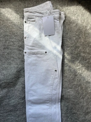 Vita jeans från Filippa K - Snygga vita jeans från Filippa K med raka ben och carpender detaljer. Jeansen har silverfärgade nitar och är tillverkade i bomull. Perfekt för dig som vill ha en clean och stilren look. Jeansen är helt oanvända och har prislappen kvar. De är i storlek 34 och har nypriset 3200kr. 
