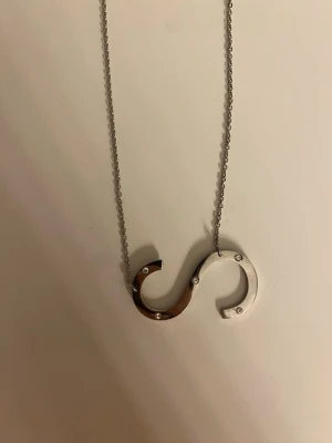 Silverfärgat halsband från IOAKU - Unikt halsband i silverfärg från IOAKU med ett S-format hänge dekorerat med små stenar. Kedjan är tunn och har justerbar längd. Perfekt för dig som gillar stilrena och moderna smycken. 