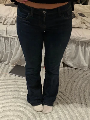 Mörkblå bootcut jeans med kontrastsömmar - Säljer ett par mörkblå bootcut jeans med snygga kontrastsömmar på bakfickorna. Klassisk femficksmodell och normal midja. Jeansen har en knapp och dragkedja framtill. Perfekta för dig som gillar en tidlös och clean look.