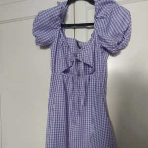 Lila rutig kortklänning H&M Divided - Superfin lila och vit rutig kortklänning från H&M Divided. Klänningen har puffärmar, knytning framtill och dragkedja i sidan. Tillverkad i lätt bomullstyg, perfekt för varma dagar. Söt och trendig modell med voluminösa ärmar och markerad midja.