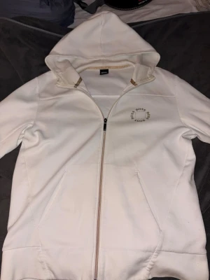 Vit hoodie från Boss  - Snygg vit hoodie från Boss med dragkedja framtill och två fickor. Klassisk huva och diskret Boss-logga på bröstet. Perfekt för en clean och stilren look. Tillverkad i mjukt material som känns skönt mot huden.