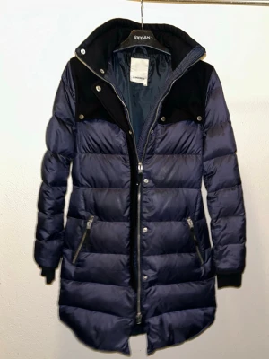 Mörkblå lång dunjacka J.Lindeberg - Hur många dunjackor får du behålla dina former ? I denna Snygga mörkblå dunjacka från J.Lindeberg med svarta detaljer vid axlar och krage har verkligen allt. Justerbara spännen i midja, stoppad med mängder av dun så den håller sig varm, många bra fickor och frsmförallt lätt att sköta om då den tål Maskintvätt och tumlare. Jackan är lång, har dragkedja, tryckknappar och två fickor med dragkedja. Perfekt för kalla vinterdagar och har en stilren look som inte går ur tiden. 