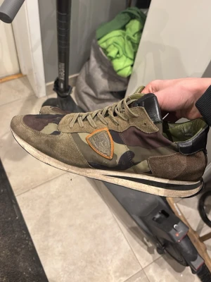  Philippe Model - Snygga sneakers från Philippe Model i camomönster med detaljer i mocka och läder. Skorna har gröna, bruna och svarta toner samt en orange logotyp på sidan. Klassisk låg modell med snörning och vit sula. Perfekta för en streetwear-look. Strl 46 nytt skick 