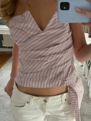 Rosa randig omlottopp med knyt - Säljer en rosa omlottopp med tunna vita och svarta ränder. Toppen har en snygg v-ringning. Ärmlös och croppad modell som ger en fräsch och trendig look. Perfekt till jeans eller kjol för en cool sommarstil.💗