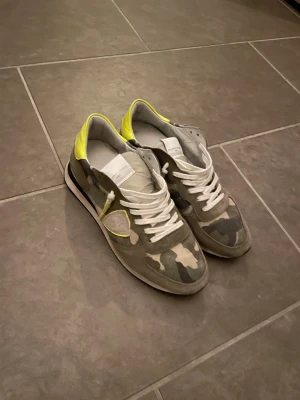 Camo sneakers med neongula detaljer - Snygga sneakers från Philippe Model med camouflagemönster i grönt, beige och grått. Skorna har vita skosnören, detaljer i mocka och coola neongula partier vid hälen. Perfekta för dig som vill sticka ut med din stil. Box m.m följer med dock lite skadad box.