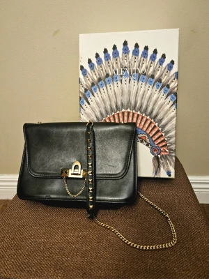 Svart Valentino Garavani Rockstud Demi-Lune Axelväska - Autentisk Valentino Garavani Rockstud Demi-Lune Axelväska – Svart & Guld  Limiterad upplaga av Valentino Rockstud Demi-Lune axelväska i svart läder med gulddetaljer. Ikonisk halvmåneformad silhuett med signaturpyramidnitar. Inkluderar original Valentino-presentask och äkthetskort. Digital kopia av originalkvitto från Valentino Saint-Tropez (09/09/2017, 1 400 €). Utmärkt skick. Tidlös lyxklassiker till ett bra pris.