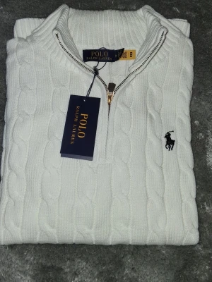 Vit quarter zip tröja Polo Ralph Lauren - Snygg vit kabelstickad tröja från Polo Ralph Lauren med halv dragkedja och klassisk broderad logga på bröstet. Tröjan har hög krage och är tillverkad i mjukt material som ger en exklusiv känsla. Perfekt för dig som gillar stilrena och tidlösa plagg.