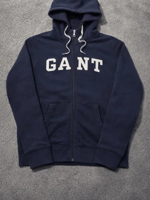 Marinblå zip hoodie från GANT - Snygg marinblå hoodie från GANT med dragkedja framtill och vit logga över bröstet. Hoodien har huva med vita dragsnören och två stora fickor framtill. Tillverkad i mjukt bomullsmaterial som känns skönt mot huden. Perfekt för en avslappnad och sportig stil.